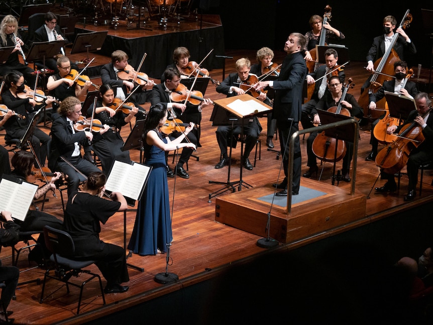 Violinist Emily Sun on stage in a blue gown performing in front of the West Australian Symphony Orchestra.