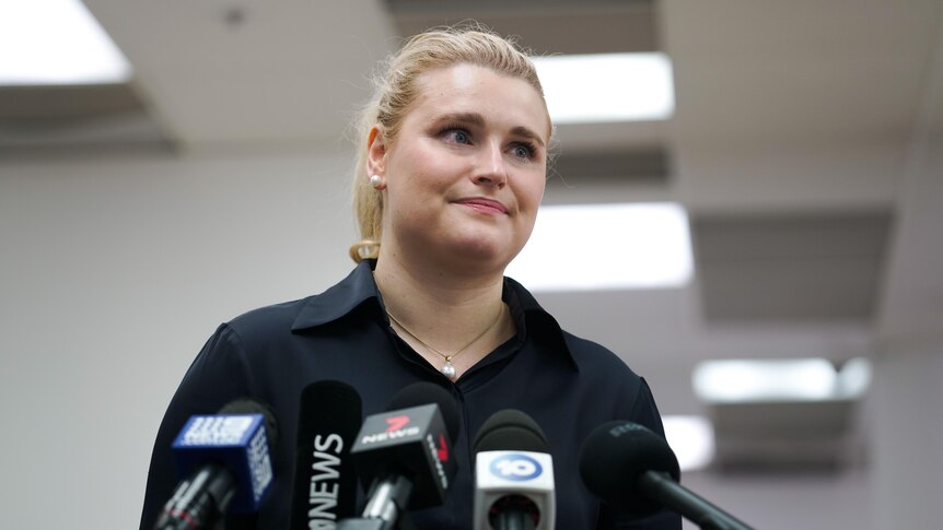 A woman with blonde hair wearing a black shirt speaks to microphones