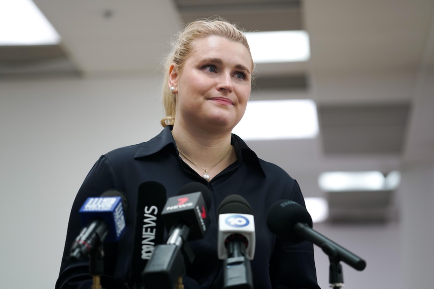 A woman with blonde hair wearing a black shirt speaks to microphones