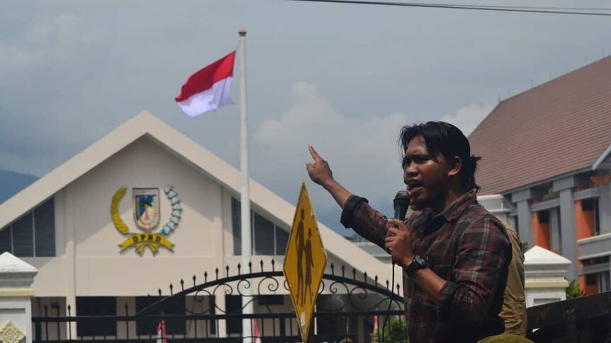 A man gestures while speaking into a microphone in front of a government office