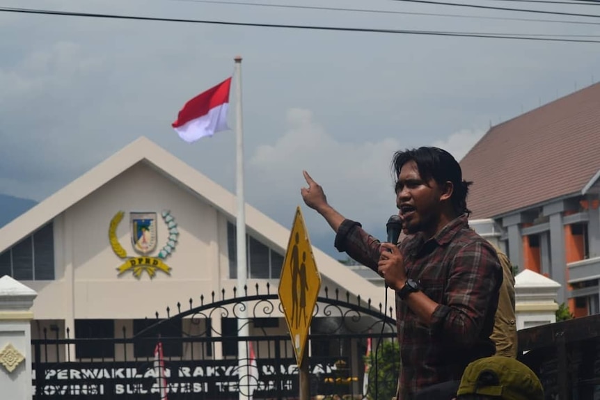 A man gestures while speaking into a microphone in front of a government office