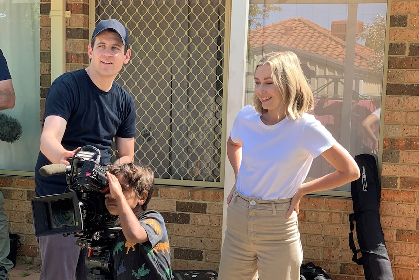 Man and woman watching a small boy looking through the viewfinder of a camera outside a house.