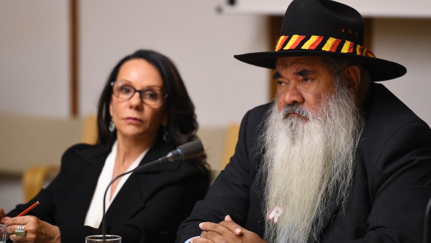 A dark-haired Aboriginal woman in a suit and glasses sits next to an older Aboriginal man with a hat, suit and long grey beard.