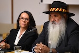 A dark-haired Aboriginal woman in a suit and glasses sits next to an older Aboriginal man with a hat, suit and long grey beard.
