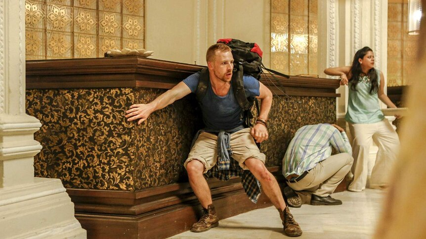 The actor crouches in fear in front of a hotel reception desk, seeking shelter from a threat out of frame.
