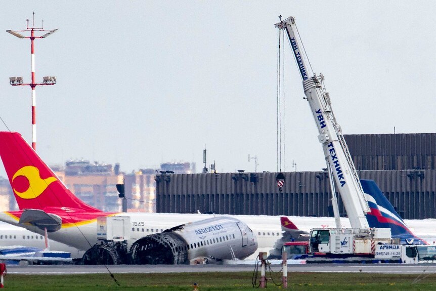 A crane lingers over a front portion of a wingless, burnt-out plane at an airport.