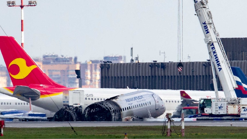 A crane lingers over a front portion of a wingless, burnt-out plane at an airport.