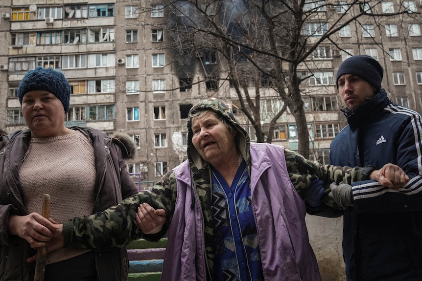 Two people help an elderly woman to walk with an apartment building hit by shelling in the background