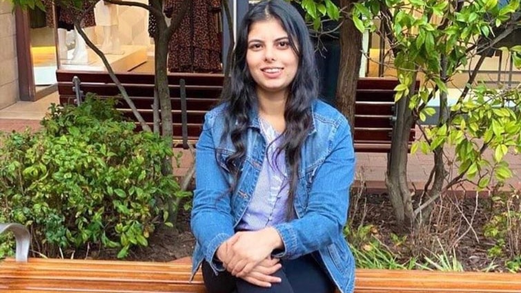 A young girl with dark black hair, wearing jeans and a denim jacket, sits cross legged and smiles at the camera.