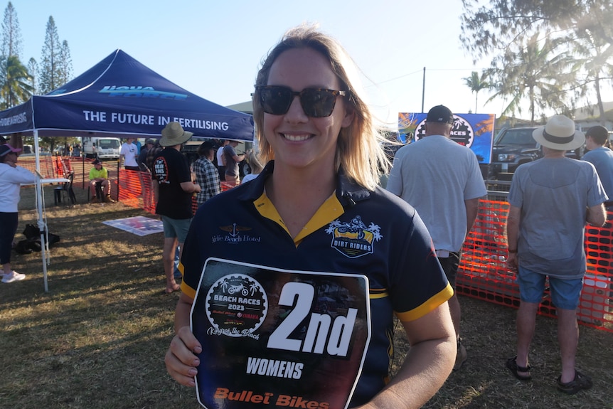 A woman wearing sunglasses and a T-shirt stands handing 2nd place sign. 