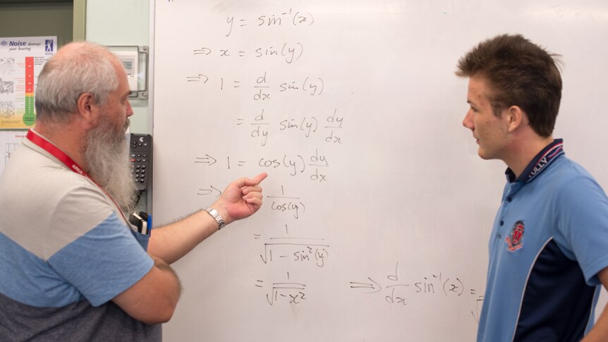 A bearded teacher talks through a complicated mathematical equation with a student.