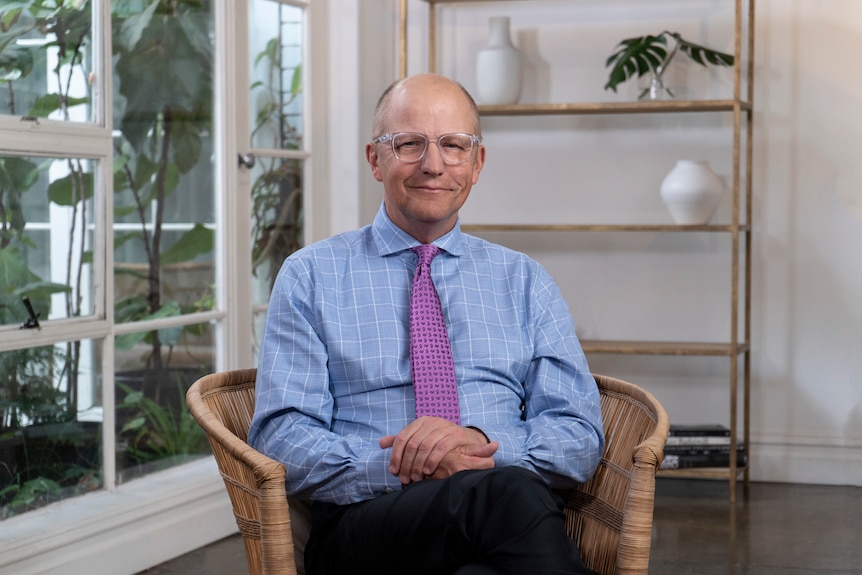 A doctor with glasses wearing a blue shirt and purple tie sitting in a chair in a nice office.