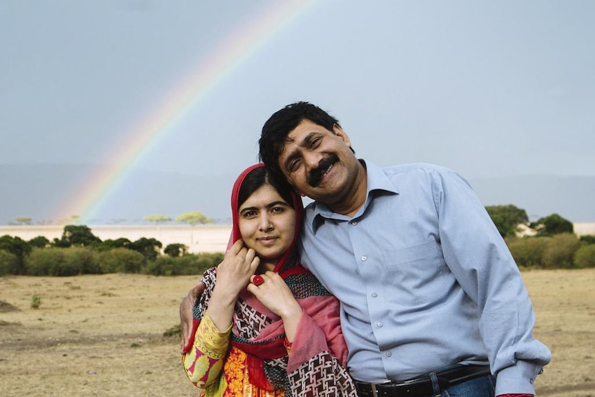 Malala Yousafzai and her father Ziauddin posing for a photo in the country with a rainbow in the background