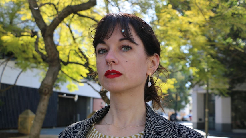 A young woman stands outside in front of a tree, looking into camera with a serious expression.