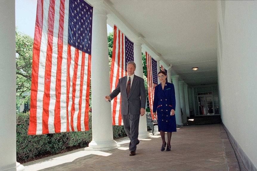 Bill Clinton and Ruth Bader Ginsburg walk along the side of the White House with american flags hung up behind them