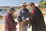 Lawyer in robes pouring sand into hands of two Indigenous men on beach with jetty in background