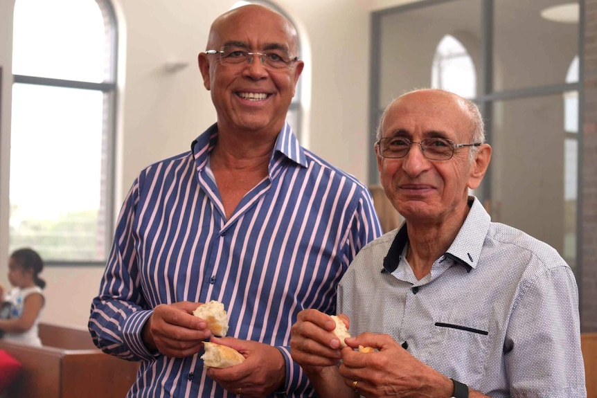 Two Egyptian men, wearing blue shirts stand in church smiling and holding bread in their hands.