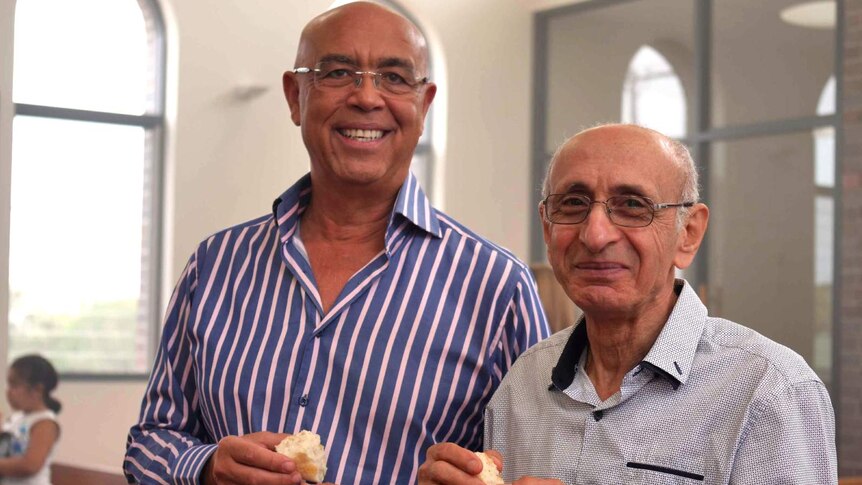 Two Egyptian men, wearing blue shirts stand in church smiling and holding bread in their hands.