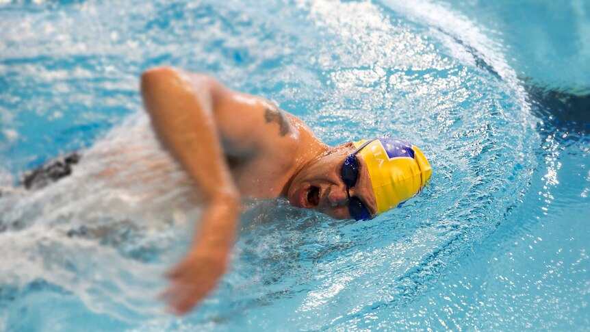 A man wearing black goggles and a yellow Australian cap comes up for air in a swimming pool