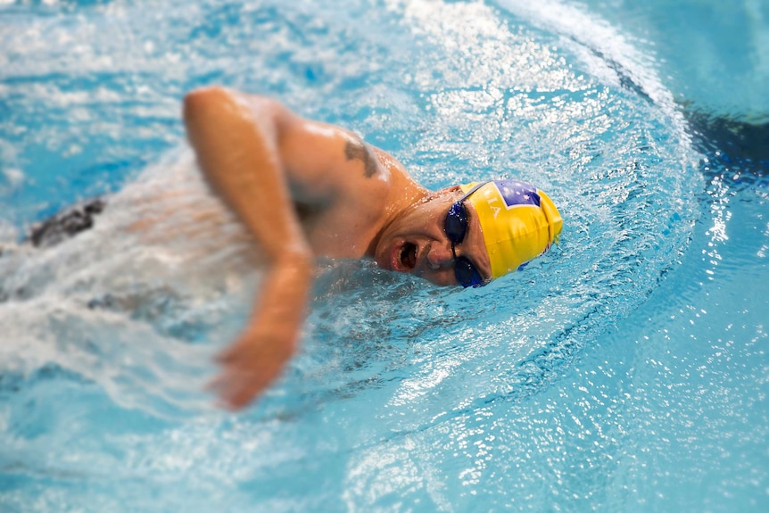 A man wearing black goggles and a yellow Australian cap comes up for air in a swimming pool