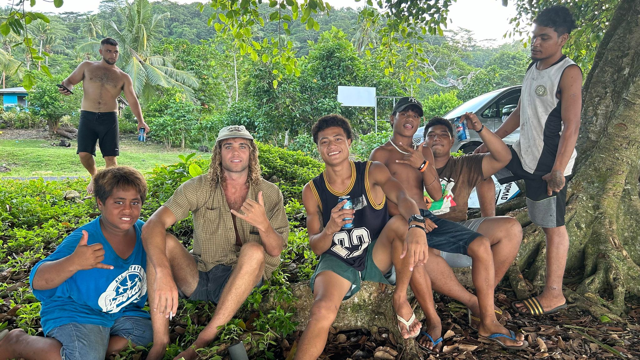 One Blonde 25 year old man 6 Island guys similar age or younger making shaka hand gestures sitting by a tree in green area 