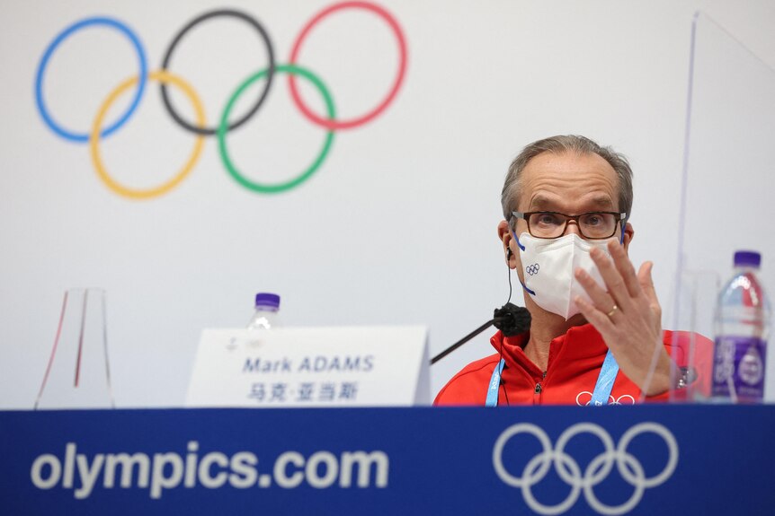 A middle-aged man in face mask and red sports shirt gestures at blue table with Olympic rings behind