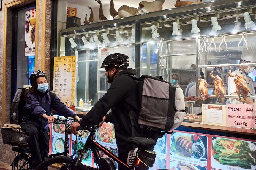 Two delivery riders on bikes wearing masks stop and talk in front of a roast duck shop