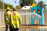 Two women wearing yellow hi-vis hold a hose spraying water at a fake blue cutout building 