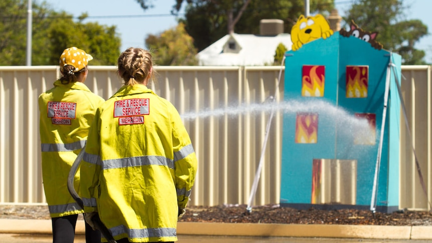 Two women wearing yellow hi-vis hold a hose spraying water at a fake blue cutout building 