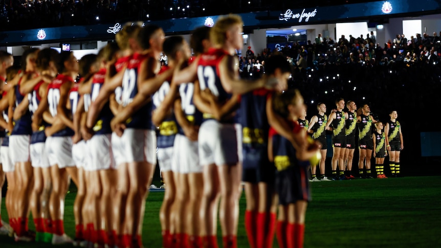 The Melbourne and Richmond AFL teams are seen standing still at attention on the MCG in a pre-match ceremony. 