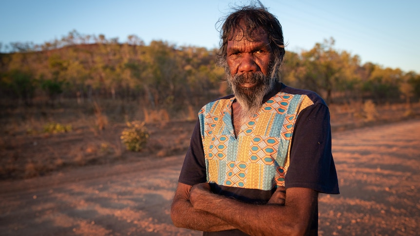 A man stands with arms crossed on a remote dirt road. 