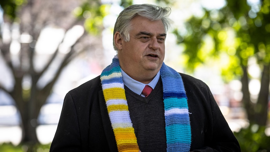 A man with a knitted scarf stands under trees.