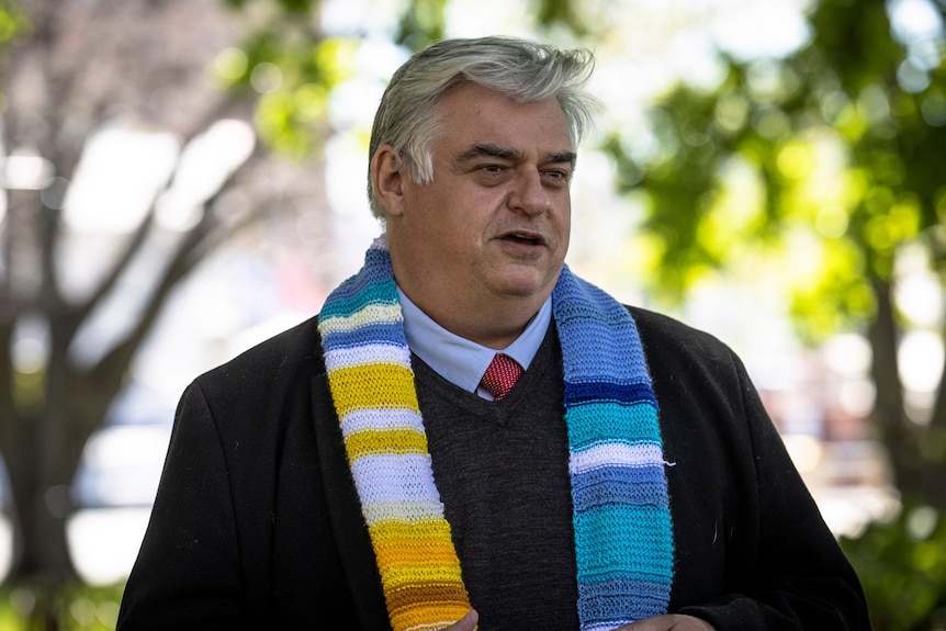 A man with a knitted scarf stands under trees.