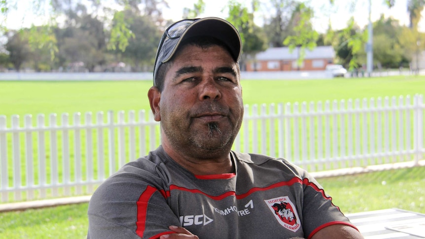 A man wearing a St George Illawarra jersey and cap sits with arms crossed.