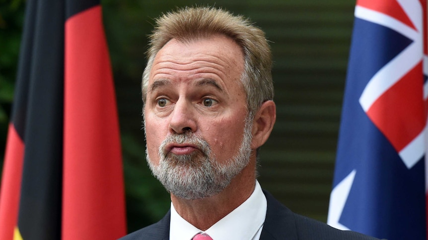 Minister for Indigenous Affairs Nigel Scullion stands in front of an Aboriginal flag and an Australian flag.