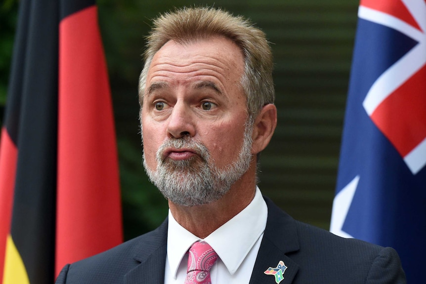 Minister for Indigenous Affairs Nigel Scullion stands in front of an Aboriginal flag and an Australian flag.
