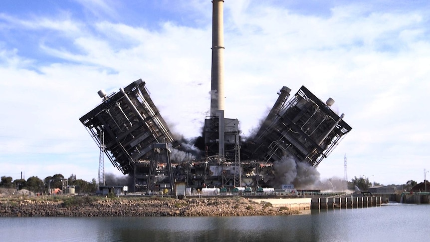 Two large structures are being demolished, falling to either side of a large power station stack.