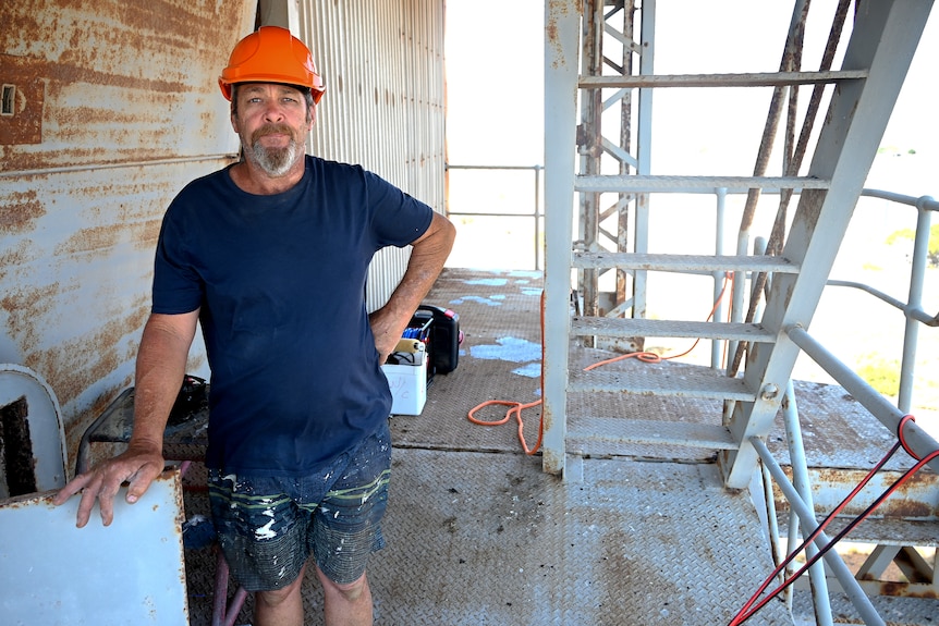 A man in an orange hard hat with a blue t-shirt stands on a deck of an old communication dish.