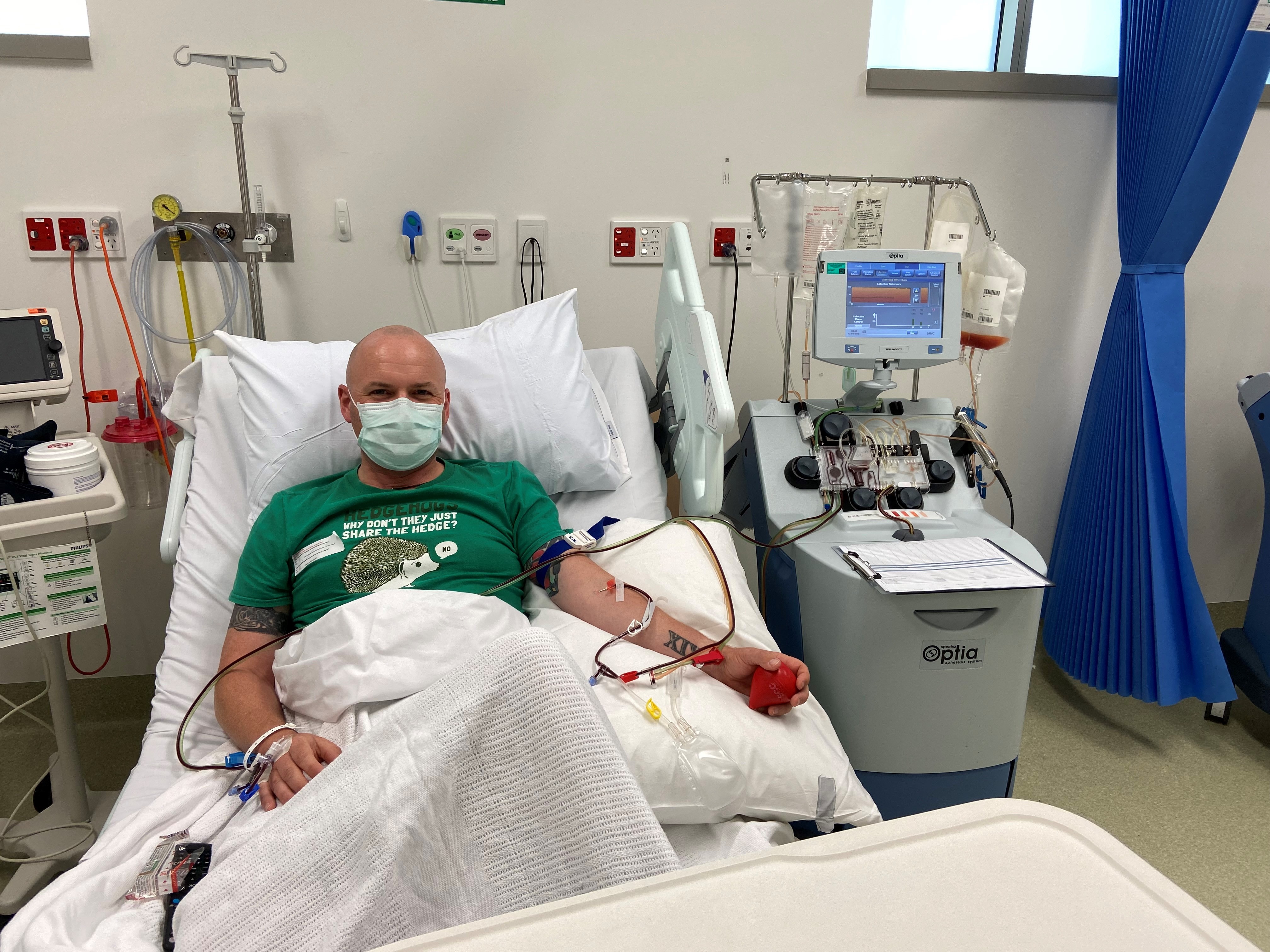 Man wearing green shirt in hospital bed with needles in arms, donating stem cells. 