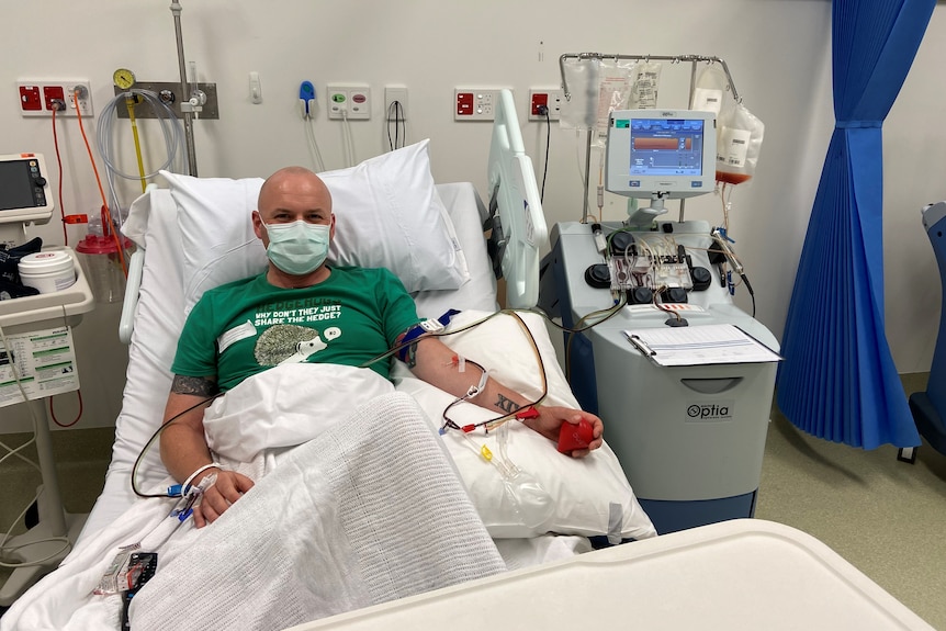 Man wearing green shirt in hospital bed with needles in arms, donating stem cells. 