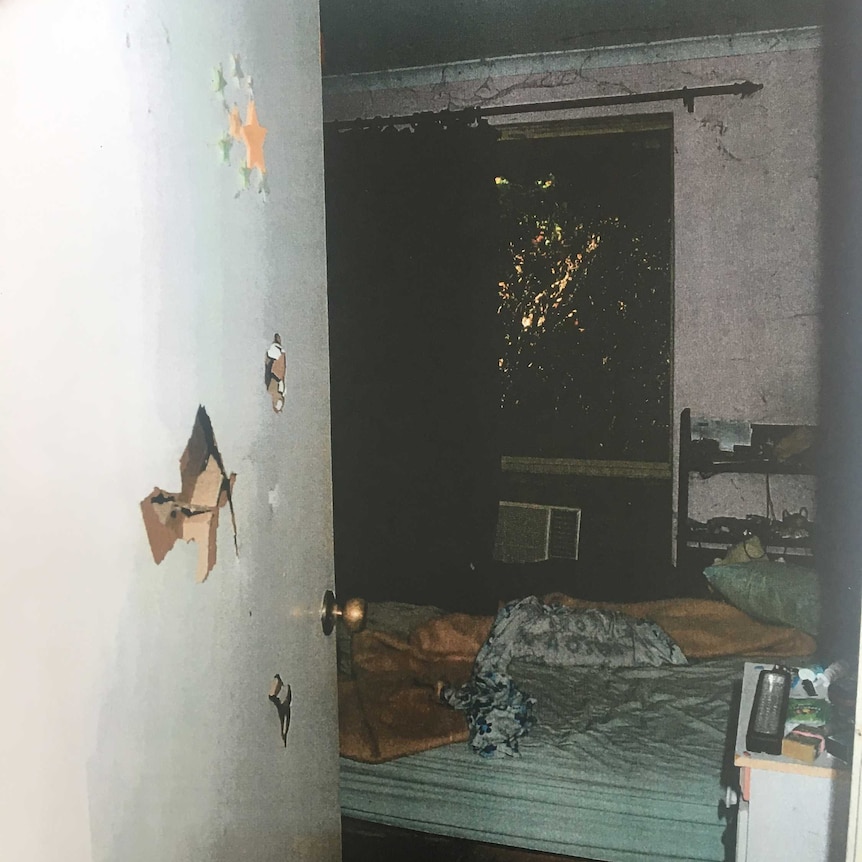 An open door with holes smashed in it leading into a dirty bedroom with an unmade bed and cobwebs above a window and curtain.
