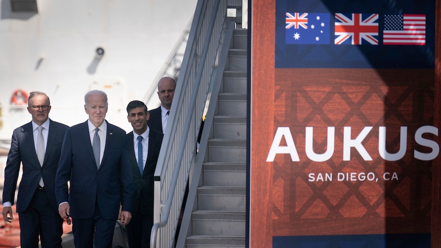 Four men walk past a flight of stars and towards a sign displaying the letters a u k u s and the flags of Australia, UK and US