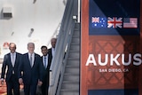 Four men walk past a flight of stars and towards a sign displaying the letters a u k u s and the flags of Australia, UK and US