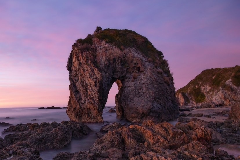An arched rock formation resembling the head and neck of a horse on a beach. The sky is pink behind the rocks. 