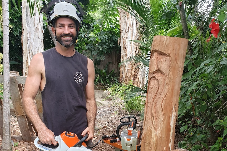 Man standing in backyard with his chainsaw and block of wood with carved face on it