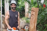 Man standing in backyard with his chainsaw and block of wood with carved face on it