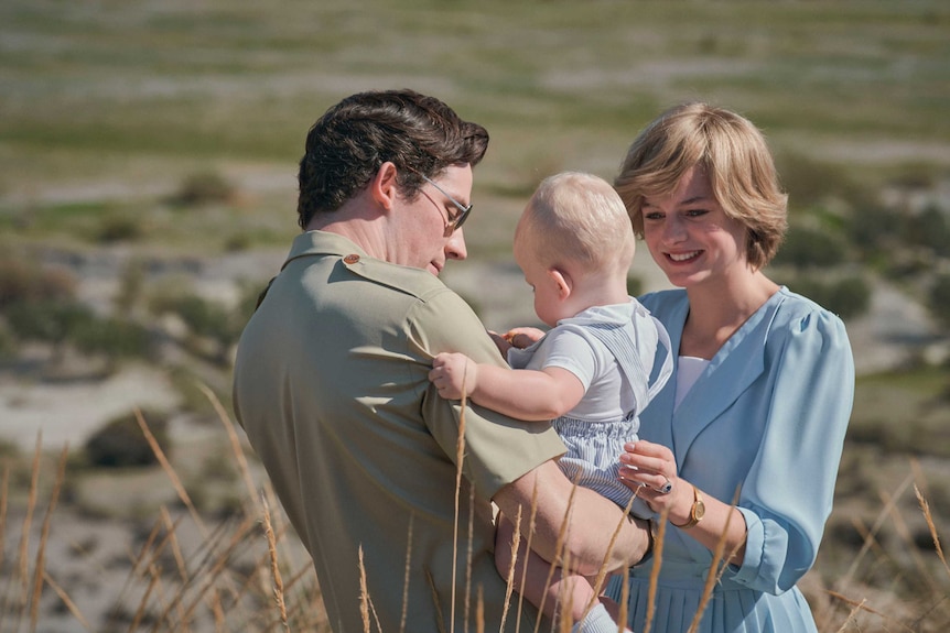 A young couple stand in an outback setting. The man is holding an infant.