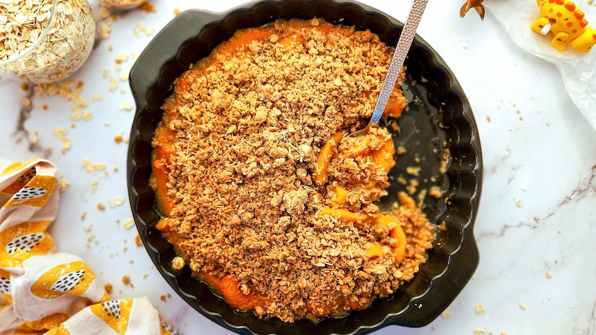 A golden-brown peach crumble in a baking dish on a white table. Oats are scattered in background.