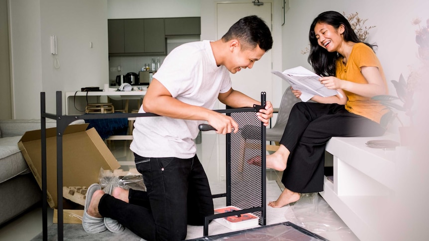 A young couple laugh as they help each other assemble a piece of flat-pack furniture.