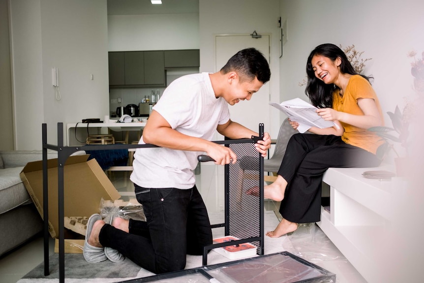 A young couple laugh as they help each other assemble a piece of flat-pack furniture.